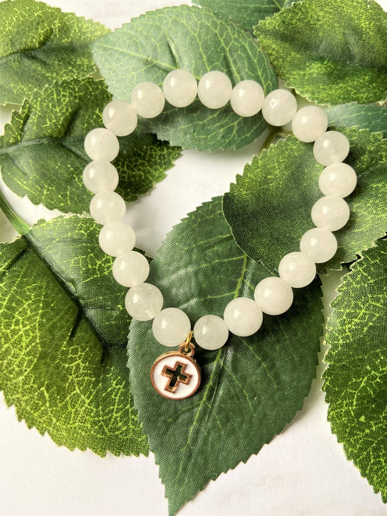 Op de afbeelding: Een armband met ronde, cr&egrave;mekleurige kralen. Een goudkleurige bedel met een witte emaille achtergrond en een zwart kruis hangt aan de armband. De armband ligt op groene bladeren.