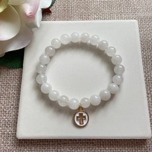 Op de afbeelding: Witte kralenarmband met een goudkleurige bedel met een wit kruis op een witte achtergrond. De armband is gemaakt van ronde, ondoorzichtige witte kralen. De bedel heeft een gouden rand.
