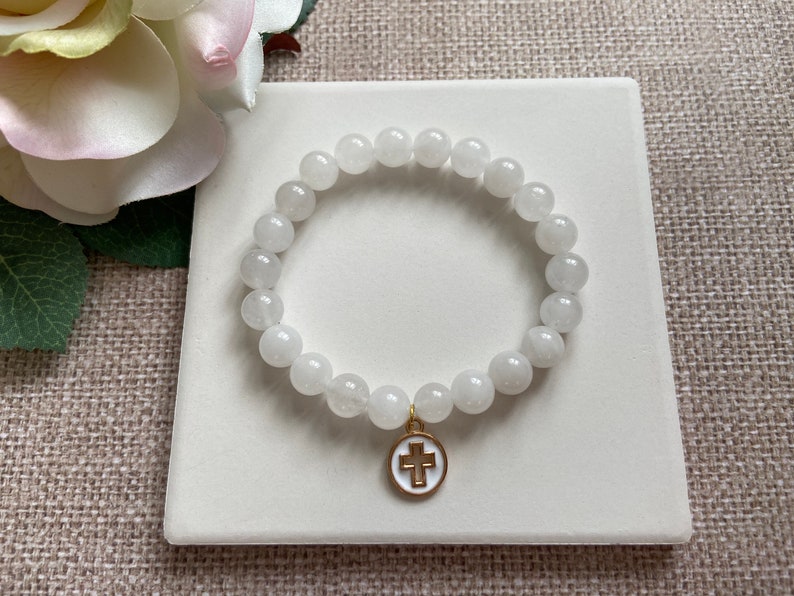 Op de afbeelding: Witte kralenarmband met een goudkleurige bedel met een wit kruis op een witte achtergrond. De armband is gemaakt van ronde, ondoorzichtige witte kralen. De bedel heeft een gouden rand.
