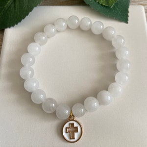 Op de afbeelding: Witte kralenarmband met een goudkleurige kruisbedel. De armband heeft ronde, ondoorzichtige witte kralen. De kruisbedel heeft een witte emaille achtergrond en een goudkleurige omtrek.