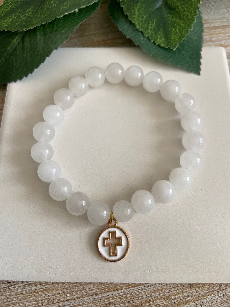 Op de afbeelding: Witte kralenarmband met een goudkleurige kruisbedel. De armband heeft ronde, ondoorzichtige witte kralen. De kruisbedel heeft een witte emaille achtergrond en een goudkleurige omtrek.