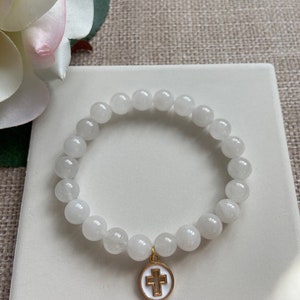Op de afbeelding: Witte kralenarmband met een goudkleurige kruisbedel. De armband is gemaakt van ronde, witte kralen en wordt gepresenteerd op een wit vierkant oppervlak. Op de achtergrond een witte en roze bloem.