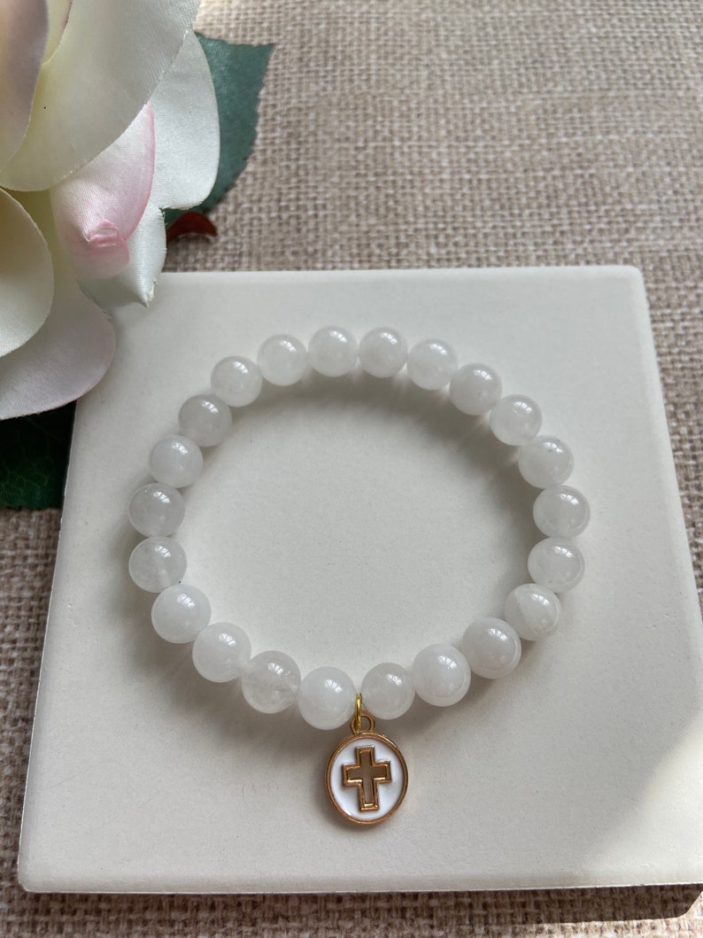 Op de afbeelding: Witte kralenarmband met een goudkleurige kruisbedel. De armband is gemaakt van ronde, witte kralen en wordt gepresenteerd op een wit vierkant oppervlak. Op de achtergrond een witte en roze bloem.