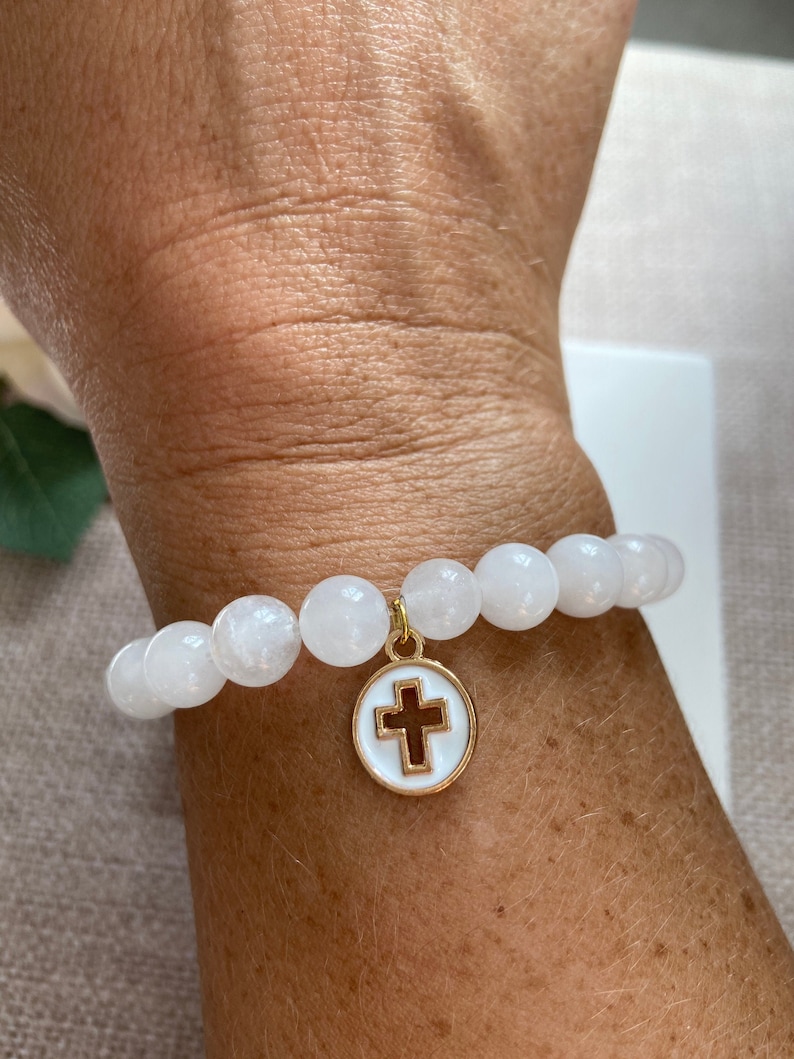 Op de afbeelding: Witte kralenarmband met een gouden bedel met een witte emaille cirkel en een bruin kruis. De armband is gemaakt van ronde, witte kralen en wordt om de pols gedragen.
