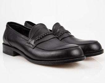 May include: A pair of black leather loafers with a braided leather strap detail. The loafers have a rounded toe and a low heel.