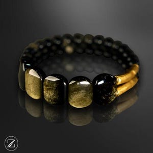Peut inclure: Un bracelet de perles noir et or. Le bracelet présente une combinaison de petites perles noires rondes et de plus grandes perles de goldstone rectangulaires. Les perles de goldstone ont un aspect chatoyant et métallique. Le bracelet est présenté sur une surface réfléchissante.