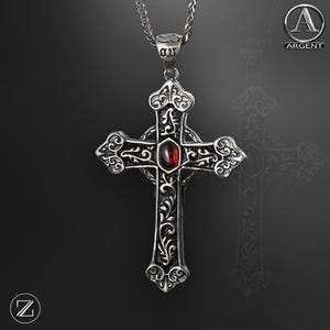 May include: A silver-toned pendant necklace featuring a detailed cross design. The cross has ornate scrollwork and a central, oval-shaped red gemstone. The necklace chain is also silver and has a textured appearance. The pendant is likely made of metal.