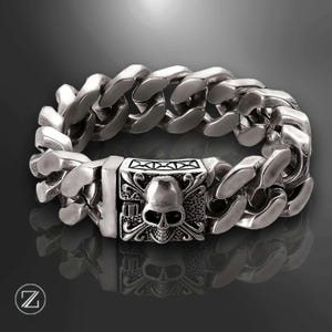 Könnte beinhalten: Ein silberfarbenes Armband mit einem Totenkopf- und gekreuzten Knochen-Design am Verschluss. Das Armband hat eine dicke Panzerketten-Optik. Der Verschluss ist rechteckig mit kunstvollen Details um den Totenkopf. Das Armband wird auf einer reflektierenden Oberfläche gezeigt.