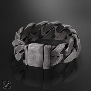 Op de afbeelding: Een armband van donkergrijs, geborsteld metaal met dikke, in elkaar grijpende schakels en een rechthoekige baksluiting. Het oppervlak van het metaal heeft overal een matte, verweerde textuur, wat zorgt voor een consistente, industriële uitstraling.