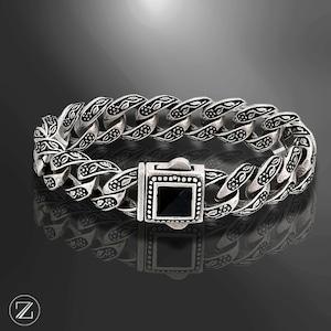 May include: Silver-toned bracelet with a curb chain design. The links feature intricate floral patterns. The clasp has a square black stone. The bracelet is displayed on a reflective surface.