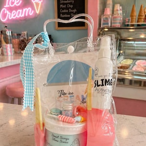 May include: A clear plastic tote bag filled with slime-making supplies. The bag contains a jar of "Cotton Candy" slime, a bottle of "Slime Activator", and various colorful accessories. The background features an ice cream shop with a neon "Ice Cream" sign.