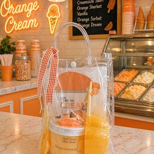 May include: A clear plastic bag filled with orange-themed items. Inside are a jar labeled "Orange Creamsicle," a small bottle, a lollipop, and a yellow spatula. The bag is tied with a red and white checkered ribbon. The background features an ice cream shop.