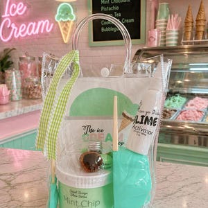 May include: A clear tote bag filled with ice cream-themed slime-making supplies. The bag contains a container of "Mint Chip" slime, a wooden spoon, a spatula, and a bottle of "Lime Activator". A green and white checkered ribbon is attached to the bag.