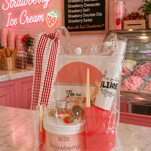 May include: A clear plastic bag contains a strawberry-themed slime kit. The kit includes a jar of "Strawberry Shortcake" slime, a pink spatula, and a bottle of slime activator. The background features a pink ice cream shop.
