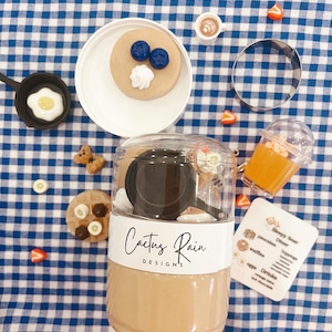 May include: A collection of miniature food items, including a pancake with blueberries and whipped cream, a fried egg, a waffle, and a cup of coffee. The items are arranged on a blue and white checkered surface, with a clear container labeled "Cactus Rain Designs."