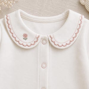 May include: A long-sleeved white shirt featuring a rounded Peter Pan collar with scalloped edges in a dusty pink thread. A small, embroidered pink rose with green leaves sits on the left side of the collar. The shirt has a front button placket.