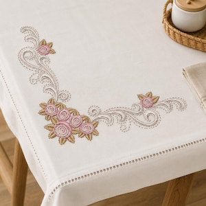 Puede incluir: Un mantel rectangular blanco con un diseño bordado en la esquina de rosas rosadas con hojas verdes y enredaderas grises. La tela tiene un detalle de borde con dobladillo calado, proporcionando un acabado limpio y estructurado al textil.