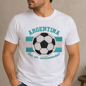 Puede incluir: Una camiseta blanca de manga corta con un diseño bordado que incluye un balón de fútbol central, una franja horizontal de rayas azul claro y blanco, y el texto "ARGENTINA" arriba y "¡Es un sentimiento!" debajo del gráfico.