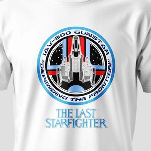 May include: White t-shirt featuring a circular graphic with a spaceship design. The graphic includes the text "IAV-500 GUNSTAR" and "DEFENDING THE FRONTIER" around the edge, with "THE LAST STARFIGHTER" below. The design is in shades of blue, red, black, and white.
