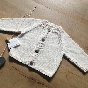 Op de afbeelding: Een wit gebreid babyvest met knopen. Het vest heeft een label met de tekst "Concept Cotton & Cashmere".