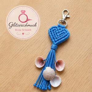 Könnte beinhalten: Ein blauer Makramee-Herz-Schlüsselanhänger mit einem silbernen Verschluss, verziert mit Muscheln. Der Schlüsselanhänger hat eine Quaste aus blauen Kordeln und ein rundes Logo mit dem Text "Glitzerschmuck Bente Schmidt".