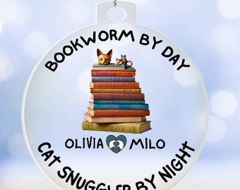 May include: White round Christmas ornament with a black and white illustration of two cats on a stack of books. The text on the ornament reads "Bookworm by Day Cat Snuggler by Night" with the names Olivia and Milo.