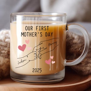 May include: A clear glass mug with a black and white illustration of two hands holding each other. The text "Our First Mother's Day" is printed on the mug, along with the names "Mommy" and "Jackson" and the year "2025".