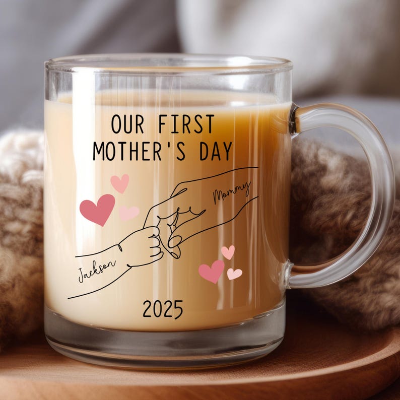 May include: A clear glass mug with a black and white illustration of two hands holding each other. The text "Our First Mother's Day" is printed on the mug, along with the names "Mommy" and "Jackson" and the year "2025".