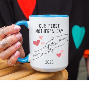 May include: White ceramic mug with a blue rim. The mug has a design of two hands holding each other, with the words "Our First Mother's Day" and the year "2025". The hands are labeled "Mommy" and "Jackson".