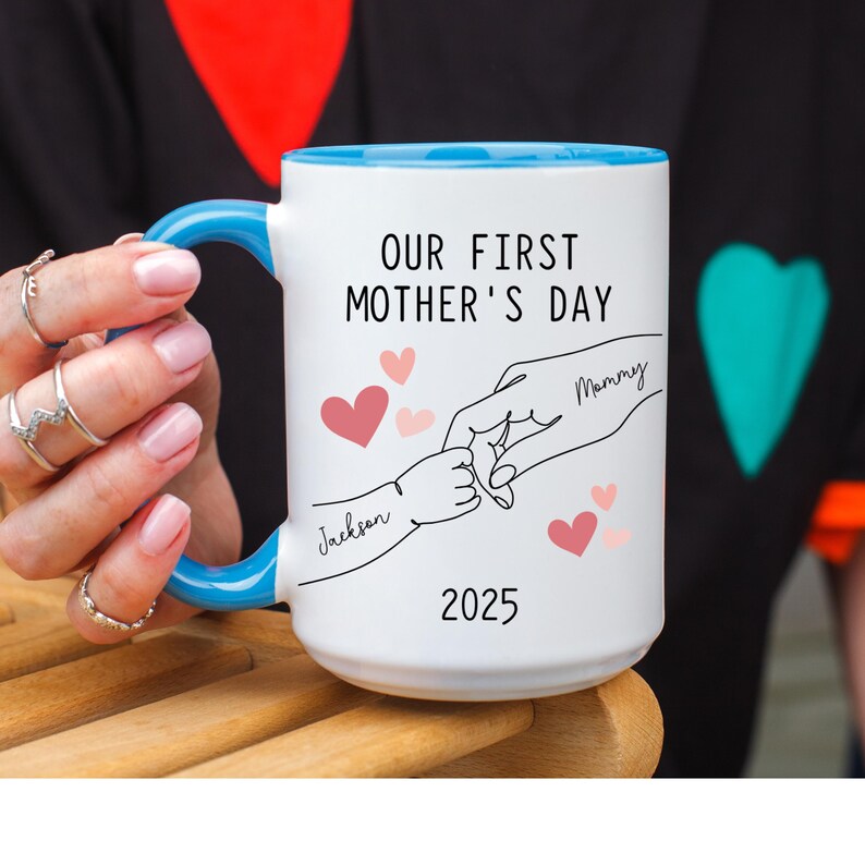May include: White ceramic mug with a blue rim. The mug has a design of two hands holding each other, with the words "Our First Mother's Day" and the year "2025". The hands are labeled "Mommy" and "Jackson".