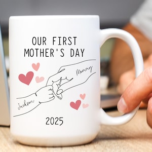 May include: White ceramic mug with black text that reads "Our First Mother's Day" and a line drawing of two hands holding each other. The text "Mommy" is written on one hand and "Jackson" is written on the other. The year "2025" is written below the hands. There are pink hearts scattered around the hands.