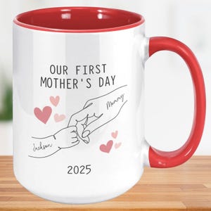 May include: White ceramic mug with a red rim and handle. The mug features a line drawing of two hands holding each other, with the text "Our First Mother's Day 2025" and the names "Mommy" and "Jackson" written on the hands.