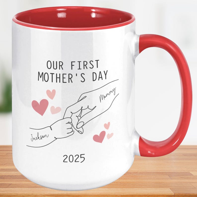 May include: White ceramic mug with a red rim and handle. The mug features a line drawing of two hands holding each other, with the text "Our First Mother's Day 2025" and the names "Mommy" and "Jackson" written on the hands.