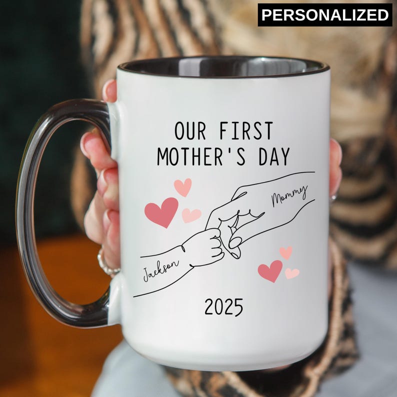 May include: White ceramic mug with a black rim. The mug has a design of two hands holding each other with the text "Our First Mother's Day" and the year "2025". The hands are labeled "Mommy" and "Jackson".