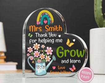 May include: A heart-shaped acrylic plaque with a black background. The plaque features a rainbow, butterflies, and a watering can with flowers. Text includes "Mrs Smith", "Thank You", "Grow and learn", and "Love Emma Wilson". A pink "SHIPS FAST" sticker is visible.