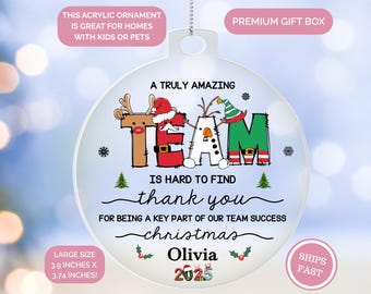 May include: A clear acrylic Christmas ornament with the word "TEAM" designed with festive characters. The ornament includes the text "A Truly Amazing", "thank you", "christmas", and the name "Olivia" with the year "2025". The ornament measures 3.9 inches x 3.74 inches.