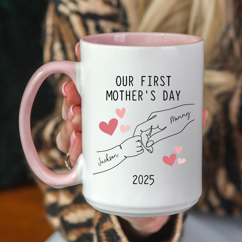 May include: White ceramic mug with a pink rim. The mug has a black outline of two hands holding each other, with the text "Our First Mother's Day" above the hands. The text "Mommy" is written on one hand and "Jackson" is written on the other. The year "2025" is written below the hands.