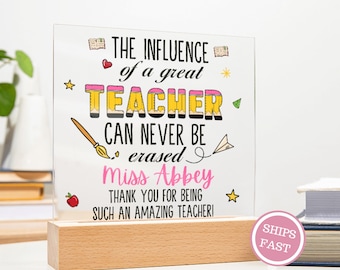 May include: A clear acrylic plaque with the text "THE INFLUENCE of a great TEACHER CAN NEVER BE erased Miss Abbey THANK YOU FOR BEING SUCH AN AMAZING TEACHER!" is mounted on a wooden base. The text is in black, pink, and yellow.