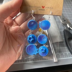 Puede incluir: Pendientes con forma de arándanos con ganchos plateados. Cada pendiente presenta tres abalorios azules, redondos y texturizados con forma de arándano. Los pendientes están sujetos a una tarjeta marrón que dice "Handmade with love."