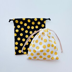 Könnte beinhalten: Zwei Kordelbeutel mit Smiley-Mustern. Ein Beutel ist schwarz mit gelben Smileys und der andere ist weiß mit gelben Smileys.