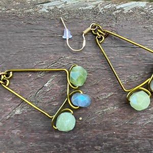 Op de afbeelding: Een paar handgemaakte goudkleurige oorbellen. Elke oorbel heeft een geometrisch driehoekig frame met drie edelsteenaccenten: groen, blauw en helder. De oorbellen worden gepresenteerd op een verweerd houten oppervlak.