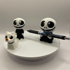 May include: Three 3D printed figurines on a white turntable. The figurines are a white ghost, a black and white skeleton with a black shirt, and a black and white skeleton with a black shirt holding a black pen with the text "ENERGEL" on it.