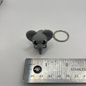 May include: A gray elephant keychain with big eyes and a textured surface. The keychain is about 1 inch tall and is attached to a silver key ring.