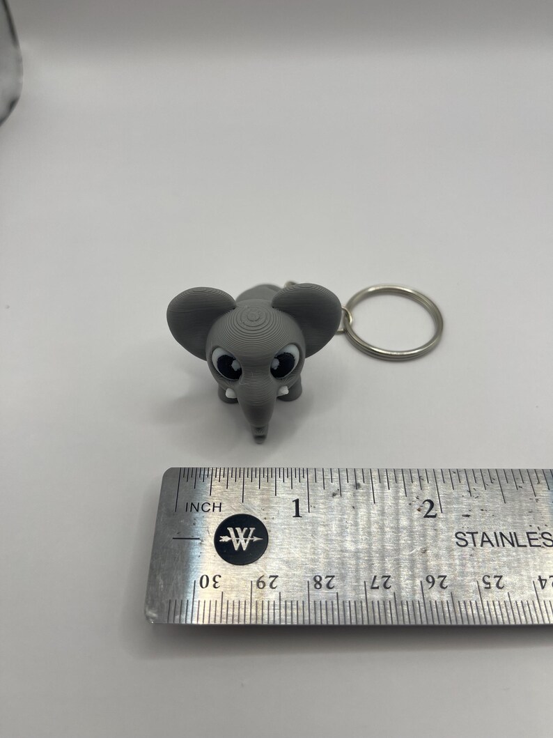 May include: A gray elephant keychain with big eyes and a textured surface. The keychain is about 1 inch tall and is attached to a silver key ring.