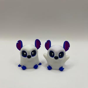 May include: Two white 3D printed figurines of small animals with blue and purple ears and large black eyes. The figurines are sitting on a white surface.