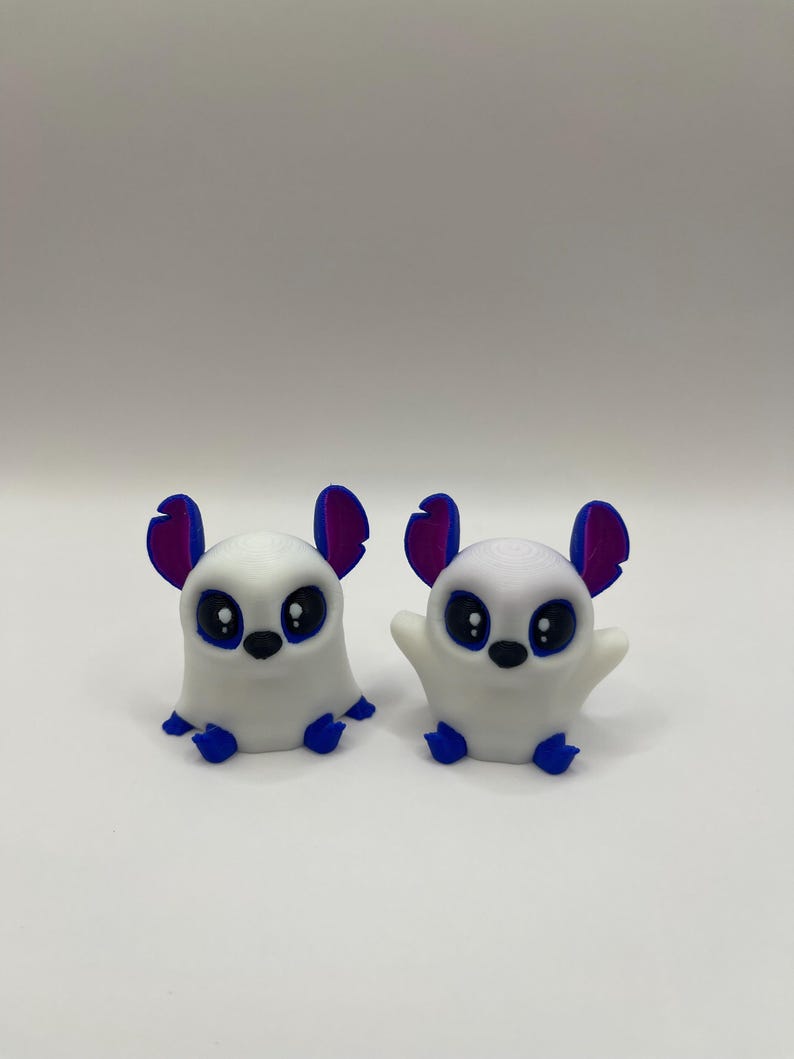 May include: Two white 3D printed figurines of small animals with blue and purple ears and large black eyes. The figurines are sitting on a white surface.