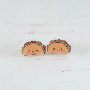 May include: Two pairs of taco-shaped earrings with smiling faces. The tacos have a light brown shell, green lettuce, and red tomato toppings. The earrings are set against a white background.
