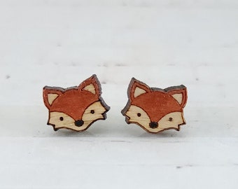 May include: A pair of wooden fox-shaped stud earrings. The earrings feature a reddish-orange fox head with a black nose and small, pointed ears. The fox's face is light-colored with black eyes. The earrings are set against a white background.