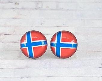 May include: A pair of round stud earrings featuring the flag of Norway. Each earring has a red background with a blue cross outlined in white. The earrings are set against a white wooden background.
