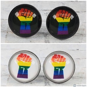 May include: A set of four round earrings. Two pairs feature a rainbow-colored fist design on a black background, and the other two have the same design on a white background. The fist is a symbol of solidarity.